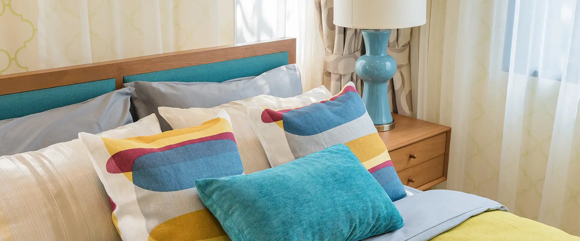 Frisch gemachtes Bett mit bunten Kissen und Bettbezug vor beige gemustertem Vorhang und Nachttisch